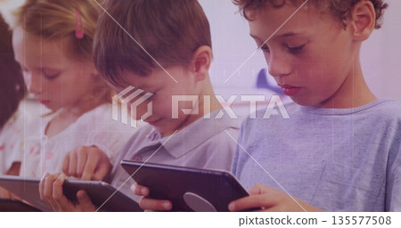 Tapping three schoolchildren using tablet devices at classroom desk, featuring posters 135577508