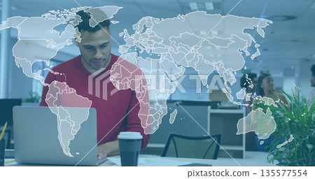 Working man wearing red sweater reviewing diagrams on laptop at open office, with world map overlay 135577554