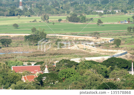 High angle view of a winding road with white walls cutting through rural green fields and forests in Thailand. Traditional temple buildings with red roofs are visible in the foreground. 135578868