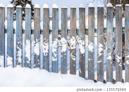 A weathered blue wooden fence stands in a snow-filled yard under a crisp winter sky A weathered blue wooden fence stands in a snow-filled yard under a crisp winter sky 135579369