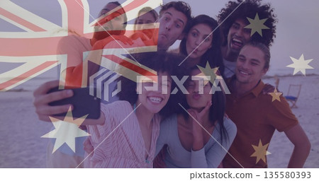 Laughing friends posing for selfie on beach at sunset with smartphone and Australian flag overlay 135580393