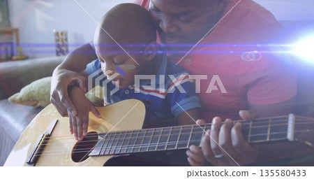 Strumming father and son playing acoustic guitar on couch at home, with decorative pillow, shelf Strumming father and son playing acoustic guitar on couch at home, with decorative pillow, shelf 135580433