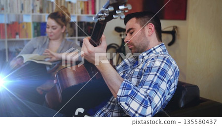 Strumming man playing acoustic guitar on sofa at home, with bicycle, woman reading book, copy space Strumming man playing acoustic guitar on sofa at home, with bicycle, woman reading book, copy space 135580437