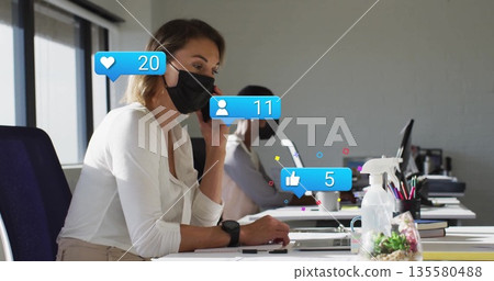 Talking on smartphone, woman wearing mask sitting in office, with laptop and social media icons Talking on smartphone, woman wearing mask sitting in office, with laptop and social media icons 135580488