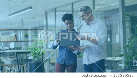 Two colleagues wearing business attire holding laptop and discussing work in open-plan office 135580583