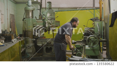 Operating machinist with safety glasses turning handwheel at shop, with milling machine, copy space Operating machinist with safety glasses turning handwheel at shop, with milling machine, copy space 135580622