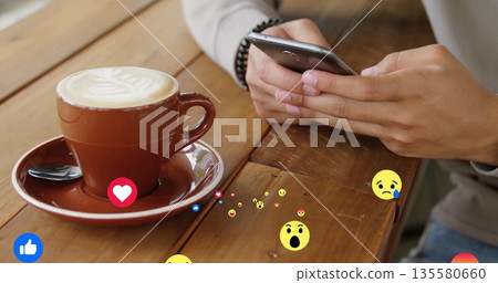 Holding smartphone tapping screen at coffee shop table, with latte cup and emoji icons, copy space 135580660