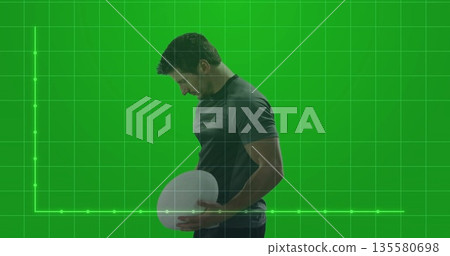 Posing male wearing dark sportswear holding white oval ball in green screen set, with grid backdrop Posing male wearing dark sportswear holding white oval ball in green screen set, with grid backdrop 135580698