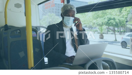 Juggling phone and laptop typing man wearing face mask and blazer on patterned bus seat 135580714