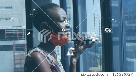 Holding smartphone masked woman giving voice command at city window, with eco icons, copy space Holding smartphone masked woman giving voice command at city window, with eco icons, copy space 135580727