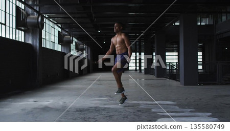 Skipping rope shirtless male exercising in industrial warehouse, wearing shorts and running shoes Skipping rope shirtless male exercising in industrial warehouse, wearing shorts and running shoes 135580749