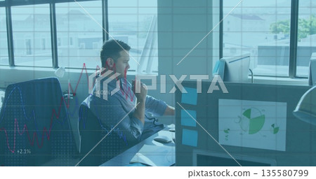 Analyzing male worker studying financial graphs on monitors at desk near windows, with sticky notes 135580799