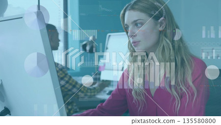 Working woman wearing pink blouse and headband focusing on desktop monitor in office, data overlays 135580800