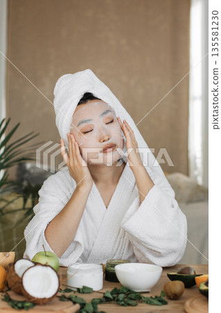 A woman with a towel on her head and wearing a bathrobe applies a facial sheet mask while surrounded by fruits and skincare products 135581230