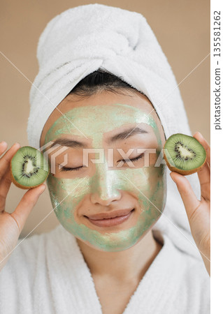 A young woman with a towel on her head and a green face mask applied to her skin holds slices of kiwi fruit near her face A young woman with a towel on her head and a green face mask applied to her skin holds slices of kiwi fruit near her face 135581262