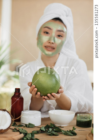 A young Asian woman with a white towel wrapped around her head and a green clay mask on her face holds a green pomelo fruit while surrounded by natural beauty products A young Asian woman with a white towel wrapped around her head and a green clay mask on her face holds a green pomelo fruit while surrounded by natural beauty products 135581273