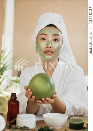 A young Asian woman with a white towel wrapped around her head and a green face mask on her face holds a green fruit while wearing a white robe A young Asian woman with a white towel wrapped around her head and a green face mask on her face holds a green fruit while wearing a white robe 135581274
