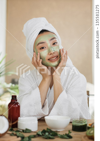 A young woman with a towel wrapped around her head applies a green facial mask while wearing a white bathrobe, surrounded by skincare products and natural ingredients 135581275
