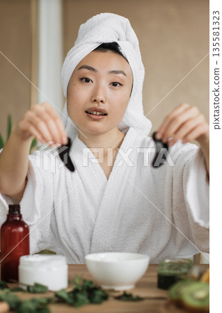 A young woman in a white bathrobe and towel wrapped around her head holds two black eye patches, ready for her skincare routine A young woman in a white bathrobe and towel wrapped around her head holds two black eye patches, ready for her skincare routine 135581323
