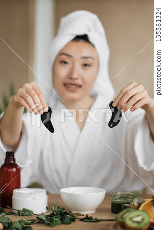 A young Asian woman wearing a white robe and towel on her head holds two black gua sha tools while preparing for a skincare routine with natural ingredients A young Asian woman wearing a white robe and towel on her head holds two black gua sha tools while preparing for a skincare routine with natural ingredients 135581324