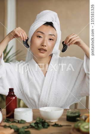 A young Asian woman in a white bathrobe and towel on her head holds two black gua sha tools near her face, surrounded by skincare products and greenery on a wooden table A young Asian woman in a white bathrobe and towel on her head holds two black gua sha tools near her face, surrounded by skincare products and greenery on a wooden table 135581326
