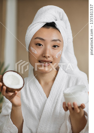 A young Asian woman with a towel on her head and wearing a white robe applies coconut cream to her face while holding a halved coconut A young Asian woman with a towel on her head and wearing a white robe applies coconut cream to her face while holding a halved coconut 135581337