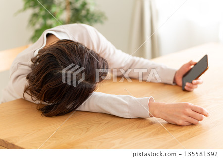 一位中年婦女懶洋洋地躺在房間裡，看著她的智慧型手機。 135581392