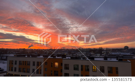 Bright colorful sunset over a city skyline with buildings and industrial pipes emitting smoke and a snow-covered landscape. Nature photography. Bright colorful sunset over a city skyline with buildings and industrial pipes emitting smoke and a snow-covered landscape. Nature photography. 135581573
