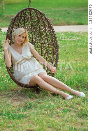 Pregnant woman in a hanging chair enjoys a moment in a grassy park on a sunny day Pregnant woman in a hanging chair enjoys a moment in a grassy park on a sunny day 135582676