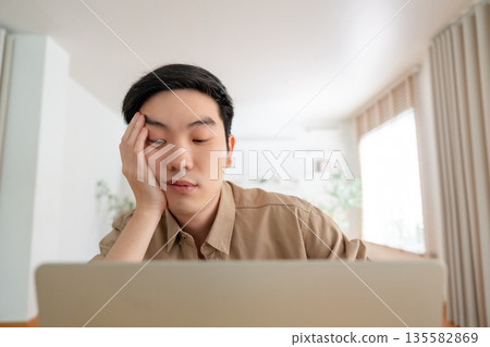 Young Asian man tired bored sad upset fatigued working on laptop in living room at home Young Asian man tired bored sad upset fatigued working on laptop in living room at home 135582869