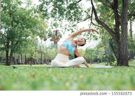 Asian woman practicing seated side stretch yoga pose outdoors in a peaceful park Asian woman practicing seated side stretch yoga pose outdoors in a peaceful park 135582873
