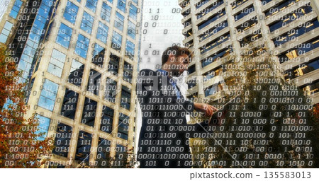 Male wearing dark suit holding phone engaging with data streams in city street, amid office towers 135583013