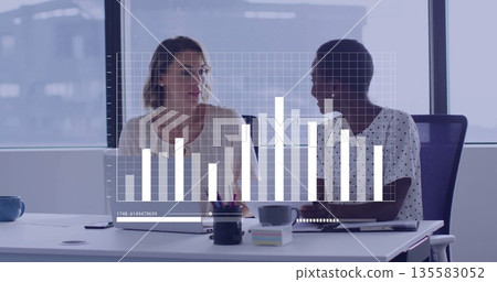Reviewing bar chart overlay by two women wearing business attire in meeting room with laptop tablet 135583052