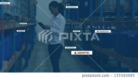 Analyzing clerk wearing uniform reviewing stock levels on tablet in warehouse aisle, with blue bins 135583095