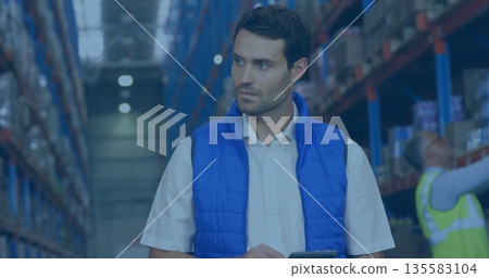 Warehouse supervisor holding tablet while inspecting pallet racks in aisle, under LED lights 135583104