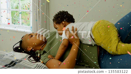 Cuddling mother in green shirt supporting baby in yellow pants on checkered blanket near window 135583122