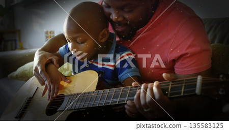 Father and son bonding over acoustic guitar on couch in living room, with cushion and bookshelf 135583125