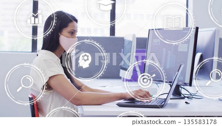 Typing Asian woman wearing mask on laptop with two monitors, floating icons in office, copy space 135583178