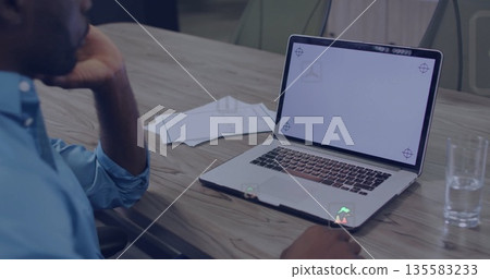 Man in blue shirt resting chin reviewing laptop in boardroom, with papers, water glass, copy space 135583233