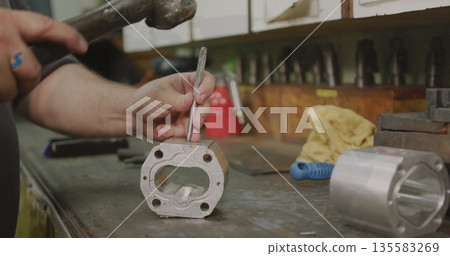 Striking machined aluminum block on metal workshop bench by worker's hands, with hammer and chisel 135583269