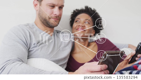 Couple sitting on couch in living room, holding tablet and smartphone with network overlay 135583294