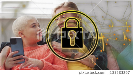 Smiling woman draping arm around companion scrolling smartphone in living room, with cushions 135583297