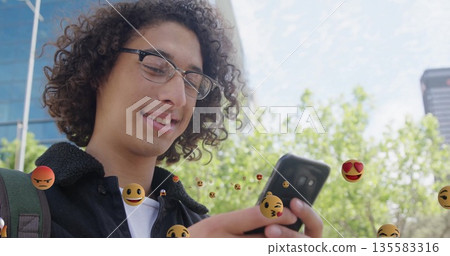 Checking student with jacket and backpack scrolling smartphone at park, floating emojis, copy space 135583316