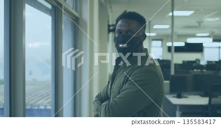 Standing man wearing olive shirt and black mask, gazing out office window with desks and monitors Standing man wearing olive shirt and black mask, gazing out office window with desks and monitors 135583417