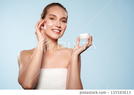 Woman applies cream to her face while holding a jar in a light blue setting Woman applies cream to her face while holding a jar in a light blue setting 135583613