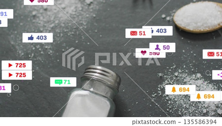 Tilting salt shaker pouring crystals across dark slate surface, with spoon and notification icons Tilting salt shaker pouring crystals across dark slate surface, with spoon and notification icons 135586394