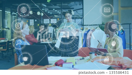 Lead designer wearing gray blazer presenting two garments in open-plan studio, with clothing rack 135586431