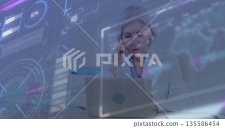 Speaking businesswoman holding smartphone while consulting laptop at office desk, with data charts Speaking businesswoman holding smartphone while consulting laptop at office desk, with data charts 135586454
