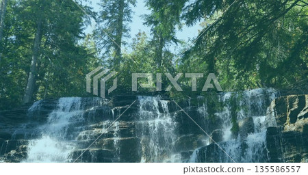 Cascading waterfall flowing over rock ledges in mountain forest, with moss patches and evergreens 135586557