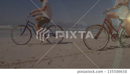 Cruising couple pedaling blue and red cruiser bikes along beach near water's edge, with ocean waves Cruising couple pedaling blue and red cruiser bikes along beach near water's edge, with ocean waves 135586638